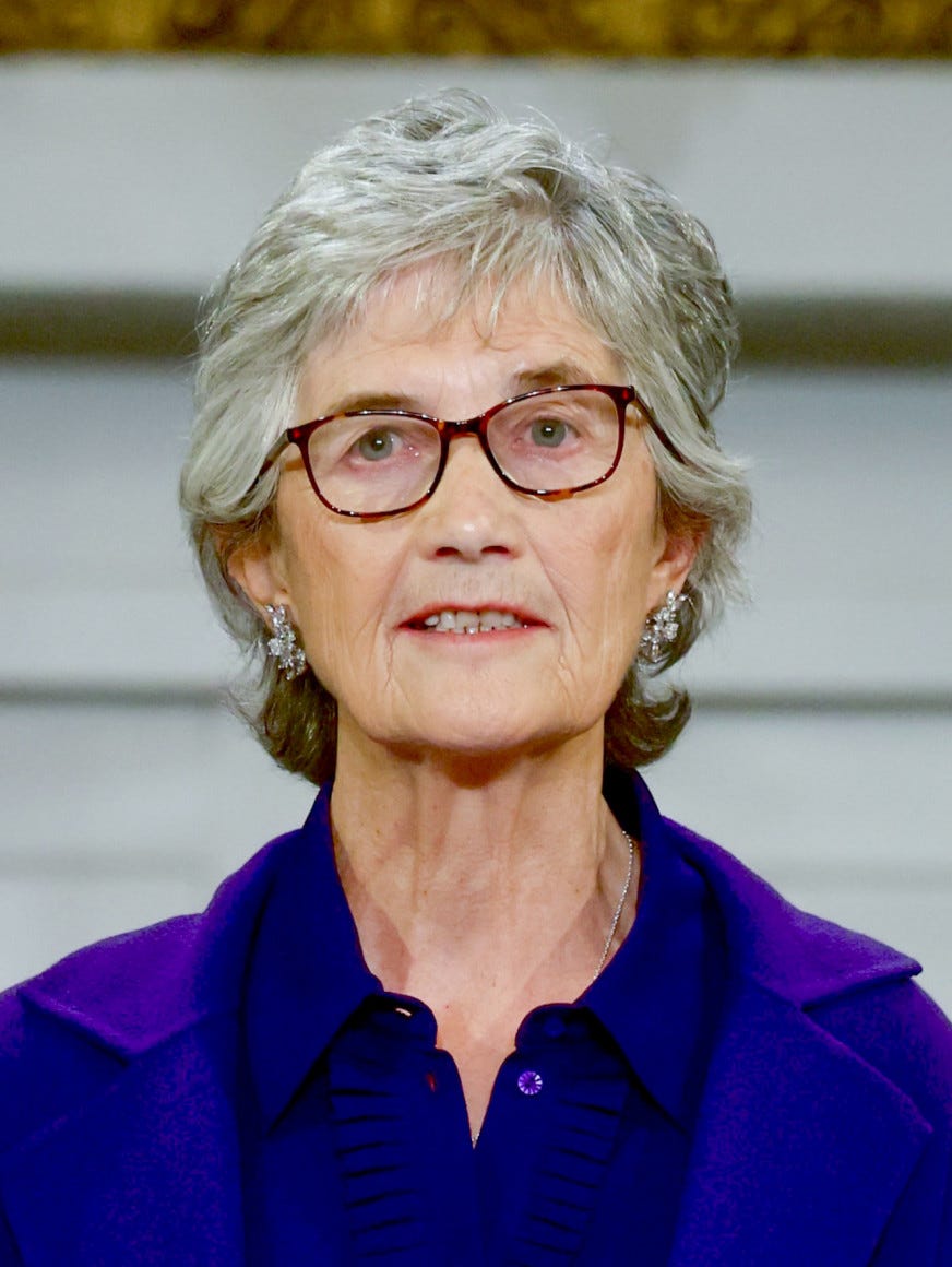 An above shoulder portrait of Catherine Connolly. She is a woman with grey and curly shoulder length hair. She is wearing a dark pinstriped blazer over a white blouse. She is smiling staring directly at the camera. An above shoulder portrait of Catherine Connolly. She is a woman with grey and curly shoulder length hair. She is wearing a dark pinstriped blazer over a white blouse. She is smiling staring directly at the camera.