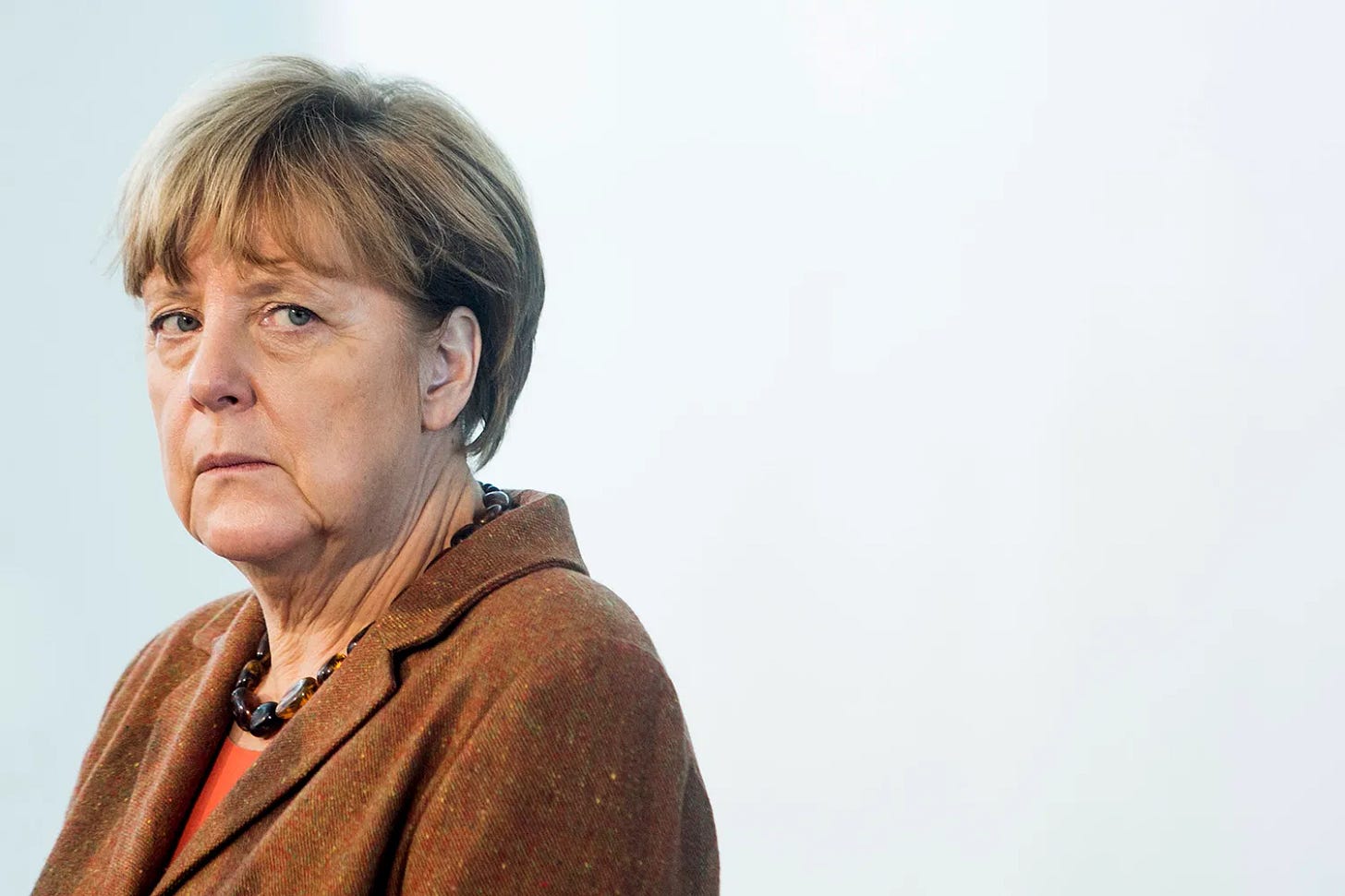 A portrait of Angela Merkel standing in front of a white wall shows Merkel from the shoulders up, wearing a serious expression and frowning slightly at something out of frame. Her hair is short and neat, and she wears a brown blazer with a chunky necklace. A portrait of Angela Merkel standing in front of a white wall shows Merkel from the shoulders up, wearing a serious expression and frowning slightly at something out of frame. Her hair is short and neat, and she wears a brown blazer with a chunky necklace.