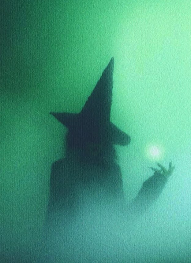 A silhouetted figure in a tall, pointed witch hat stands in thick green fog, holding up one hand toward a small glowing orb of light.