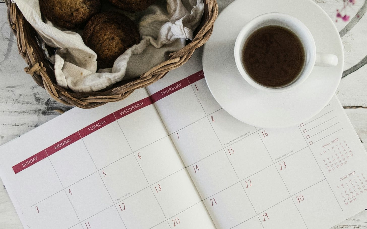 An open monthly planner with a blank calendar grid on a white table, next to a cup of black coffee and a wicker basket of baked goods. An open monthly planner with a blank calendar grid on a white table, next to a cup of black coffee and a wicker basket of baked goods.