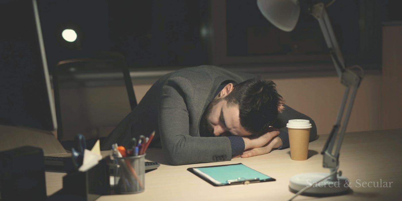 A tired man asleep at his desk late at night, surrounded by a laptop and scattered papers, illuminated by soft warm light in a quiet room.