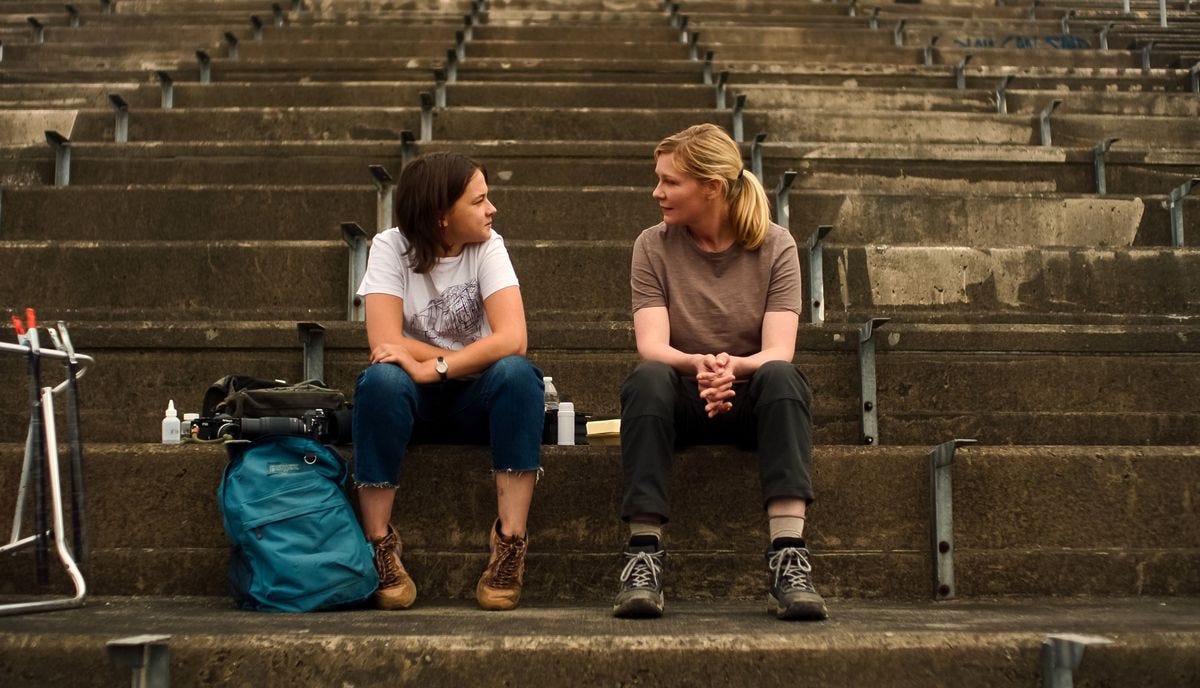 Photojournalists Lee (Kirsten Dunst) and Jessie (Cailee Spaeny) sit together on stone bleachers, surrounded by their cameras and gear, and face each other in Alex Garland’s Civil War