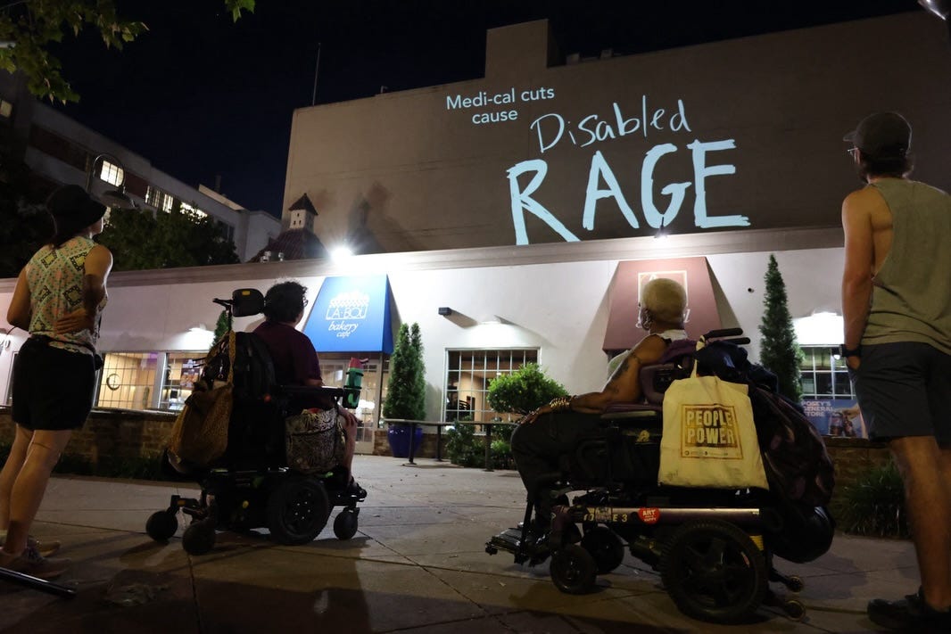 Evening photo of a projected message lit up on the front of a building that reads: Medi-Cal cuts cause DISABLED RAGE, with two people in power wheelchairs and two people standing behind them, looking up at the message. Evening photo of a projected message lit up on the front of a building that reads: Medi-Cal cuts cause DISABLED RAGE, with two people in power wheelchairs and two people standing behind them, looking up at the message.