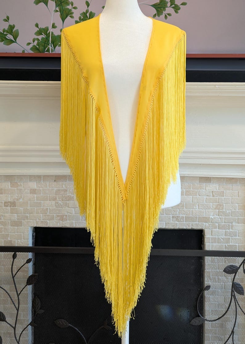 May include: A bright yellow fringed shawl displayed on a mannequin. The shawl has a V-neck design with long, flowing fringe. The garment is made of a lightweight material, and the fringe cascades down the front. The background includes a fireplace and decorative elements.