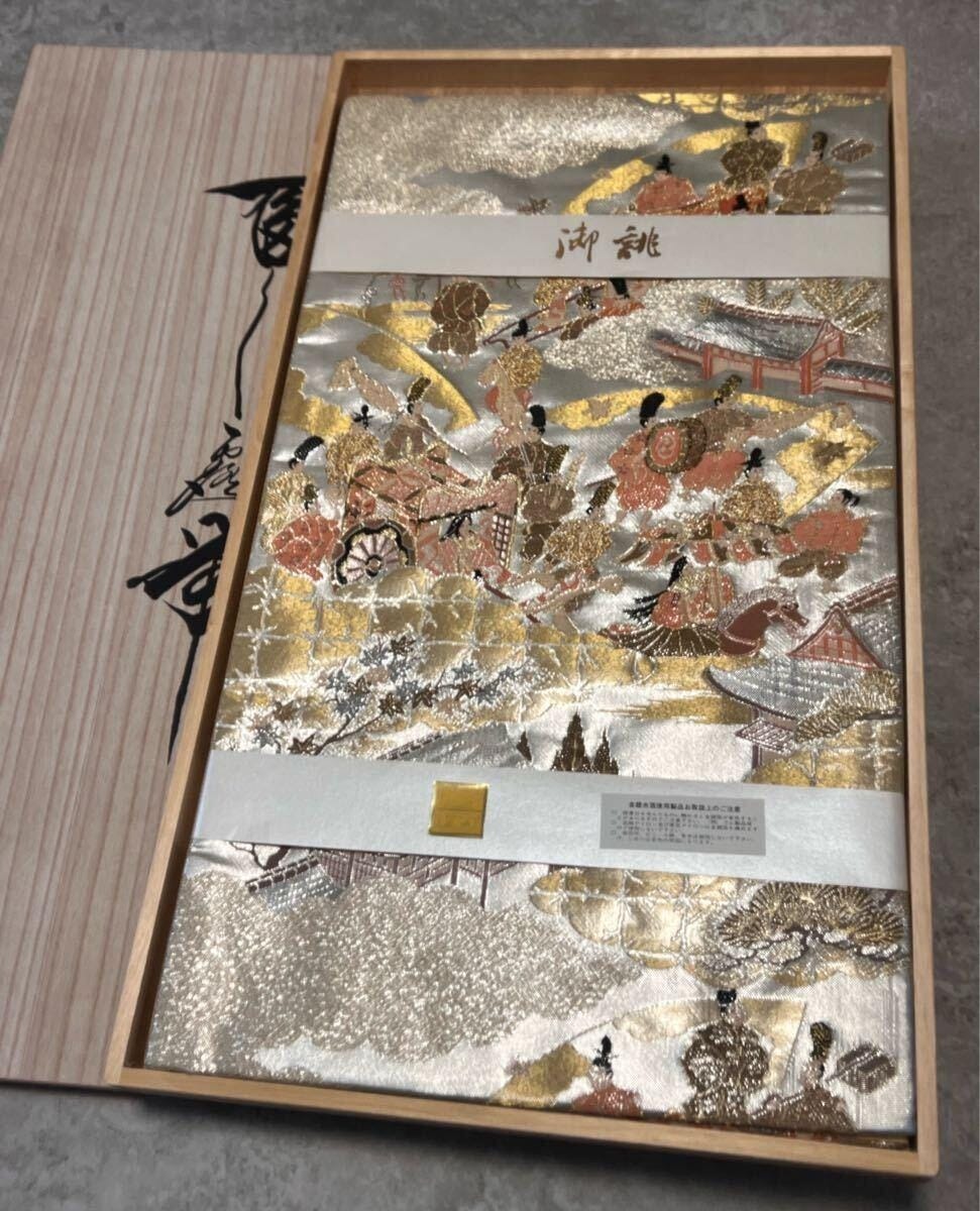 Kinran gold brocade in extreme close-up: the characteristic shimmer of gold foil wound around a silk core, catching workshop light at shifting angles. The black ground throws the metallic thread into relief — karakusa arabesques and stylised floral medallions rendered not as surface decoration but as structure built thread by thread. Kinran gold brocade in extreme close-up: the characteristic shimmer of gold foil wound around a silk core, catching workshop light at shifting angles. The black ground throws the metallic thread into relief — karakusa arabesques and stylised floral medallions rendered not as surface decoration but as structure built thread by thread.