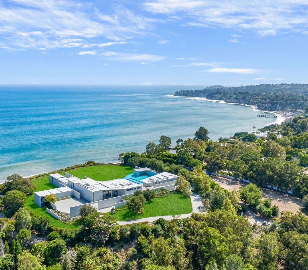 A birds-eye view of a white, sprawling mansion with a pool. It is on a green, tree-filled hillside overlooking the ocean. A birds-eye view of a white, sprawling mansion with a pool. It is on a green, tree-filled hillside overlooking the ocean.