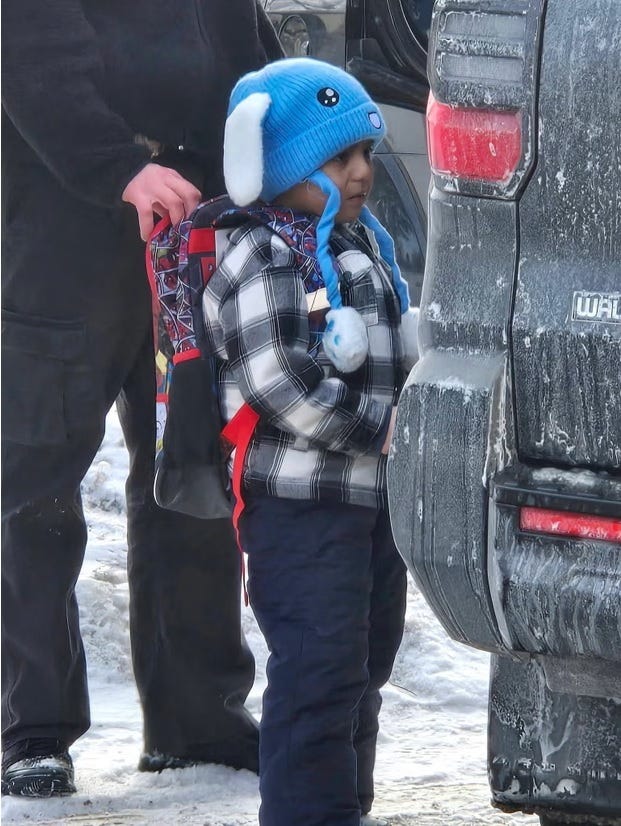 Photo of a five-year-old boy bravely trying not to cry as armed ICE goons hold him by his backpack at the side of an unmarked SUV. The boy wears snow pants, a grey plaid winter coat, and a cheerful blue and white stocking cap with an animal face on it and floppy puppy ears. 