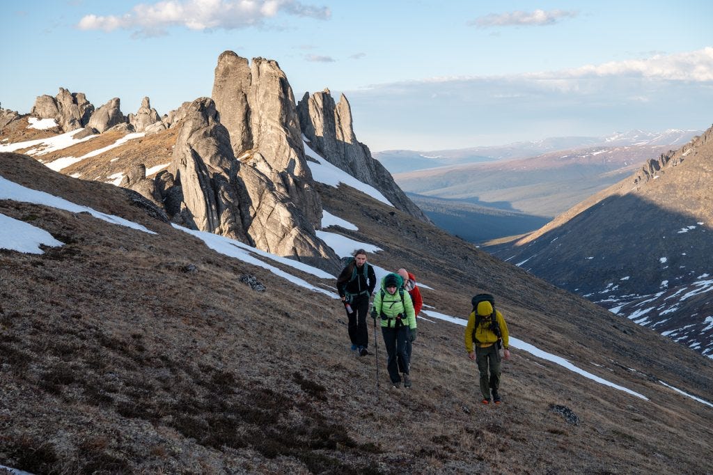 Four hikers with packs climb a brown tundra slope in low evening light, a row of tall granite spires rising along the ridge to their left and a deep valley opening to distant ridgelines on the right.
