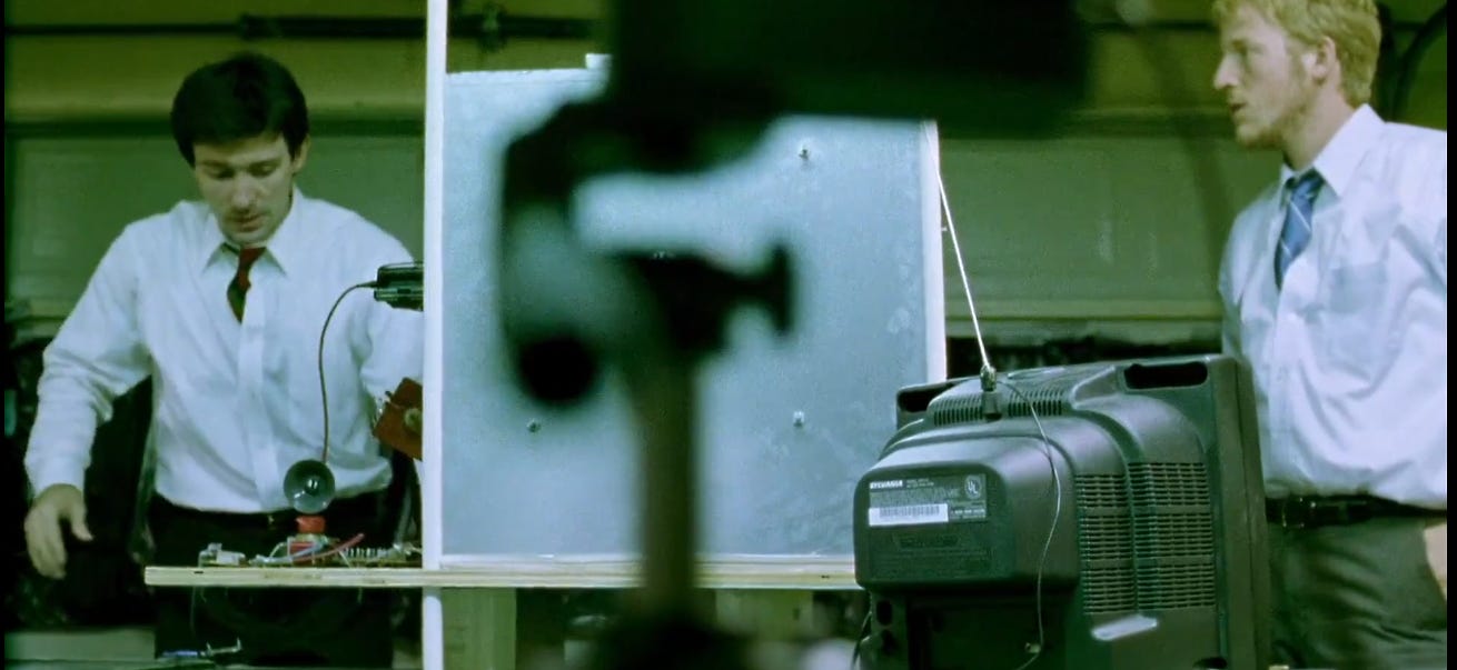 Aaron and Abe, divided by the machine they built together. The box stays centered. They drift.