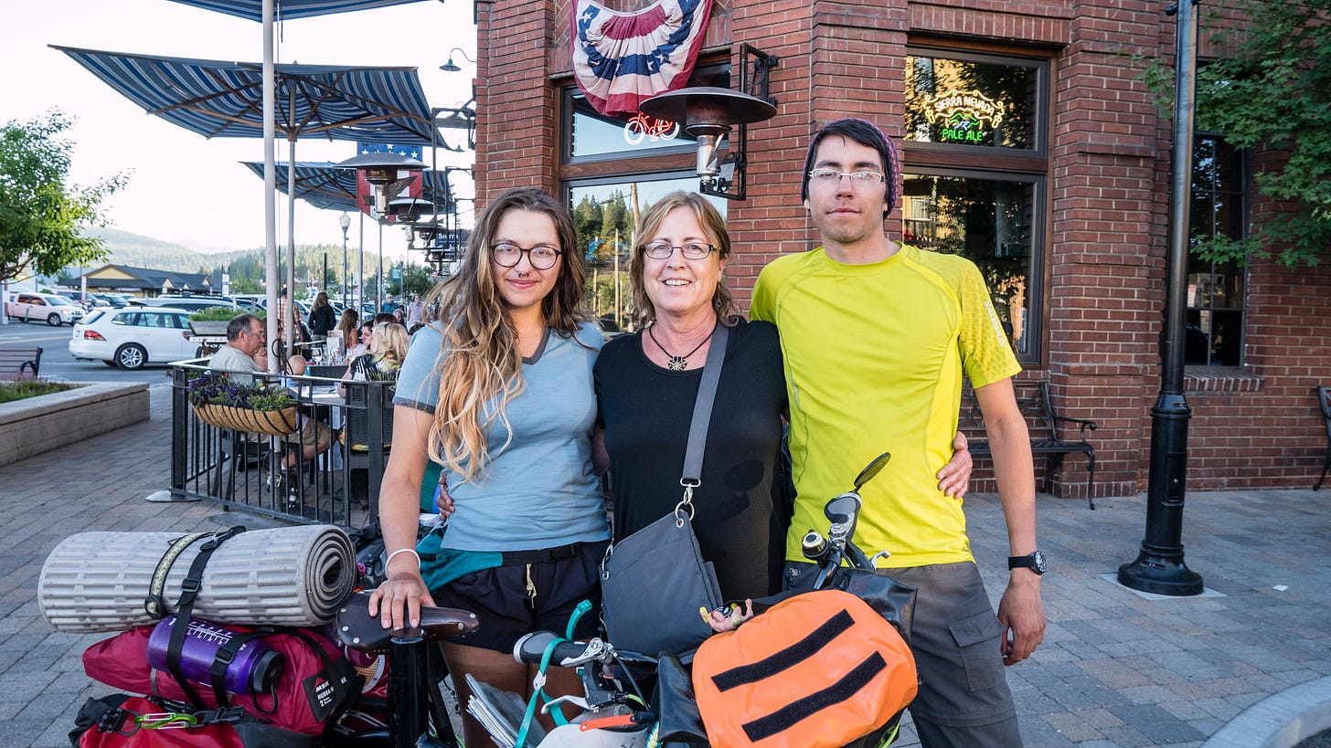 A pair of young bicyclists stand behind a loaded touring bicycle. Between the two siblings is their older aunt. The touring bicycle has bags, water bottles, and a sleeping pad rolled up and bungeed to the back. In the background is the brick facade and entrance of a restaurant with red, white, and blue bunting over the door. Tables of people are sitting outside the restaurant, eating.