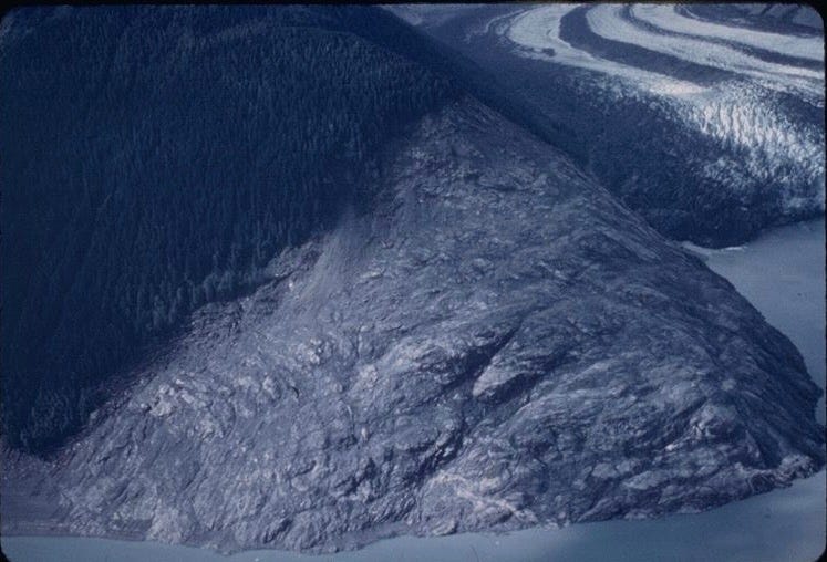 A rocky headland, stripped bare of vegetation high on its slope, rises from a body of water. In the background, a glacier terminates in the water. A rocky headland, stripped bare of vegetation high on its slope, rises from a body of water. In the background, a glacier terminates in the water.