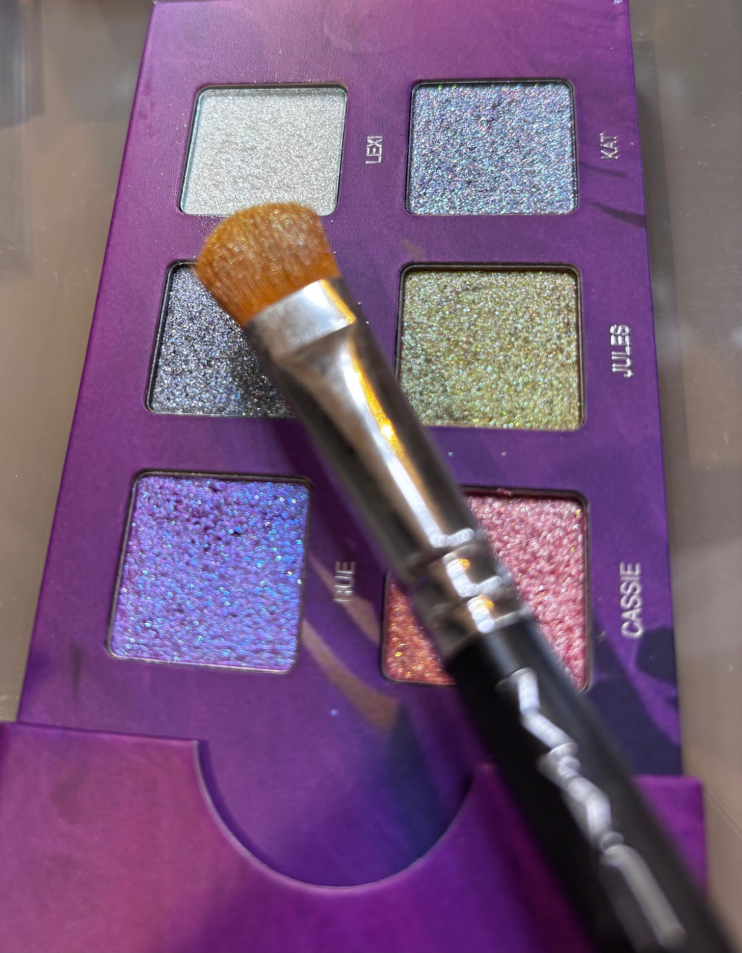 A macro shot of the Half Magic x Euphoria eyeshadow palette with a M.A.C makeup brush resting on the highly pigmented, glittery pans.