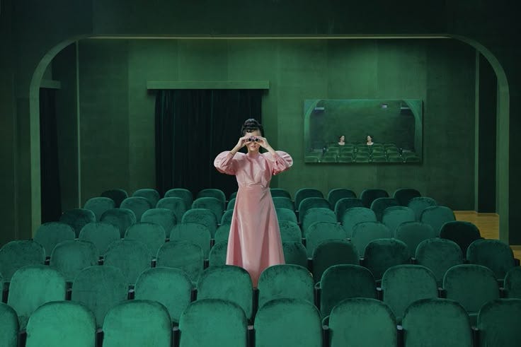 This may contain: a woman in a pink dress standing in front of a green auditorium filled with chairs
