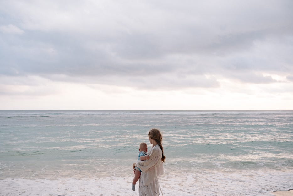 A mother stands beside her child on a quiet beach at sunset, symbolizing love, guidance, and the gentle strength of motherhood.
