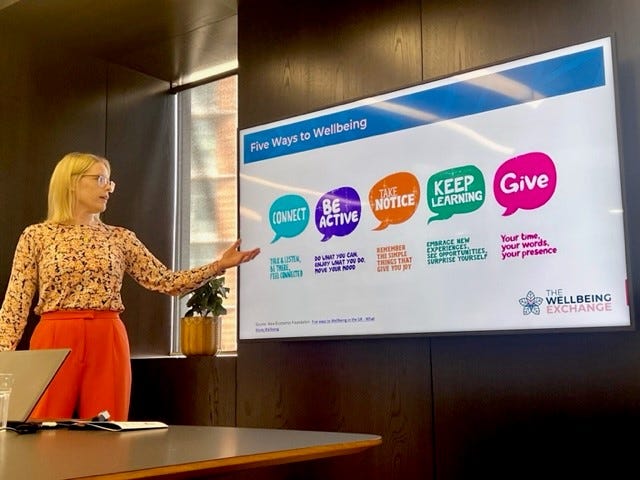 A woman in orange trousers with blond hair and glasses, stands behind a table with a laptop and points to a presentation screen. The slide on the screen has the Five Ways to Wellbeing on it and the logo The Wellbeing Exchange. 