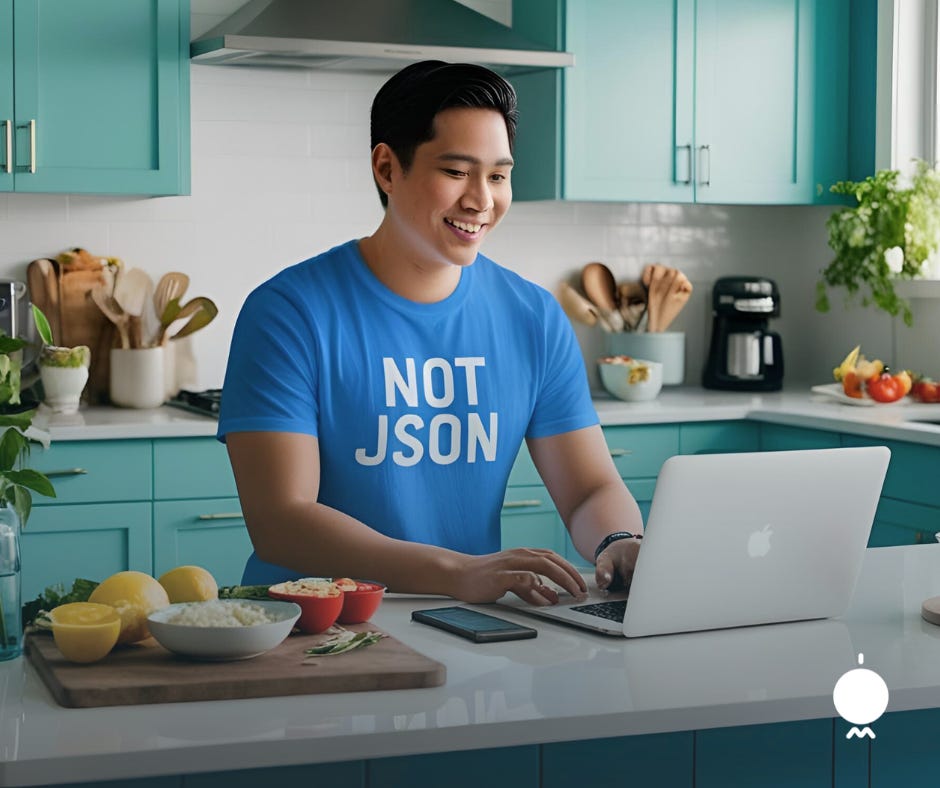 A smiling man in a bright blue kitchen wearing a shirt that says “NOT JSON” as he types on a laptop. Fresh ingredients are on the counter beside him, suggesting he’s working on a recipe. A smiling man in a bright blue kitchen wearing a shirt that says “NOT JSON” as he types on a laptop. Fresh ingredients are on the counter beside him, suggesting he’s working on a recipe.