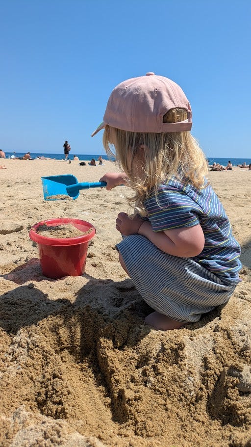 Blonde toddler filling a bucket with sand on a sunny beach, she is looking away from the camera and wearing a pink cap