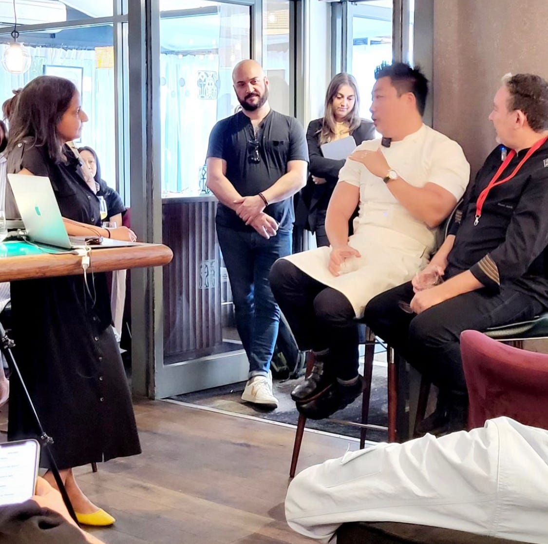Three people, two sitting, one standing engaged in a discussion while others look on in the dining area of a restaurant Three people, two sitting, one standing engaged in a discussion while others look on in the dining area of a restaurant