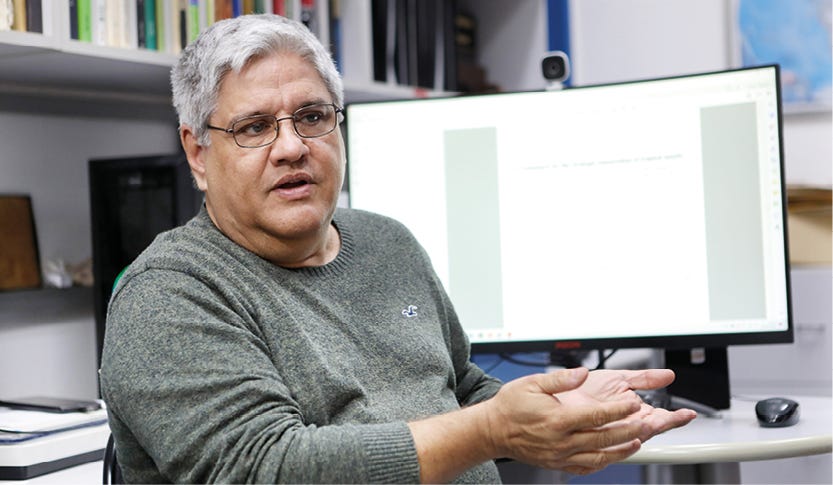 Pessoa de cabelos grisalhos usando óculos e suéter cinza claro, sentada em ambiente de escritório com estantes ao fundo, gesticulando com a mão direita aberta enquanto olha para a esquerda, com monitor de computador exibindo documento em branco posicionado à sua direita. Pessoa de cabelos grisalhos usando óculos e suéter cinza claro, sentada em ambiente de escritório com estantes ao fundo, gesticulando com a mão direita aberta enquanto olha para a esquerda, com monitor de computador exibindo documento em branco posicionado à sua direita.