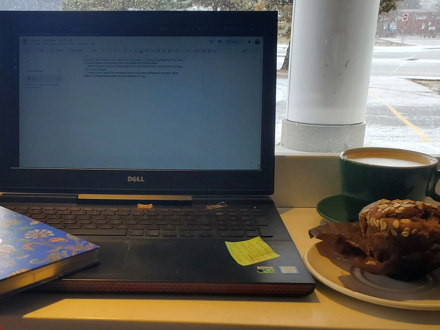 A laptop sits on a bar table looking out at a parking lot. The corner of a notebook peeks into view at the bottom left corner of the keyboard. A latte sits behind a muffin on a saucer to the right of the laptop.