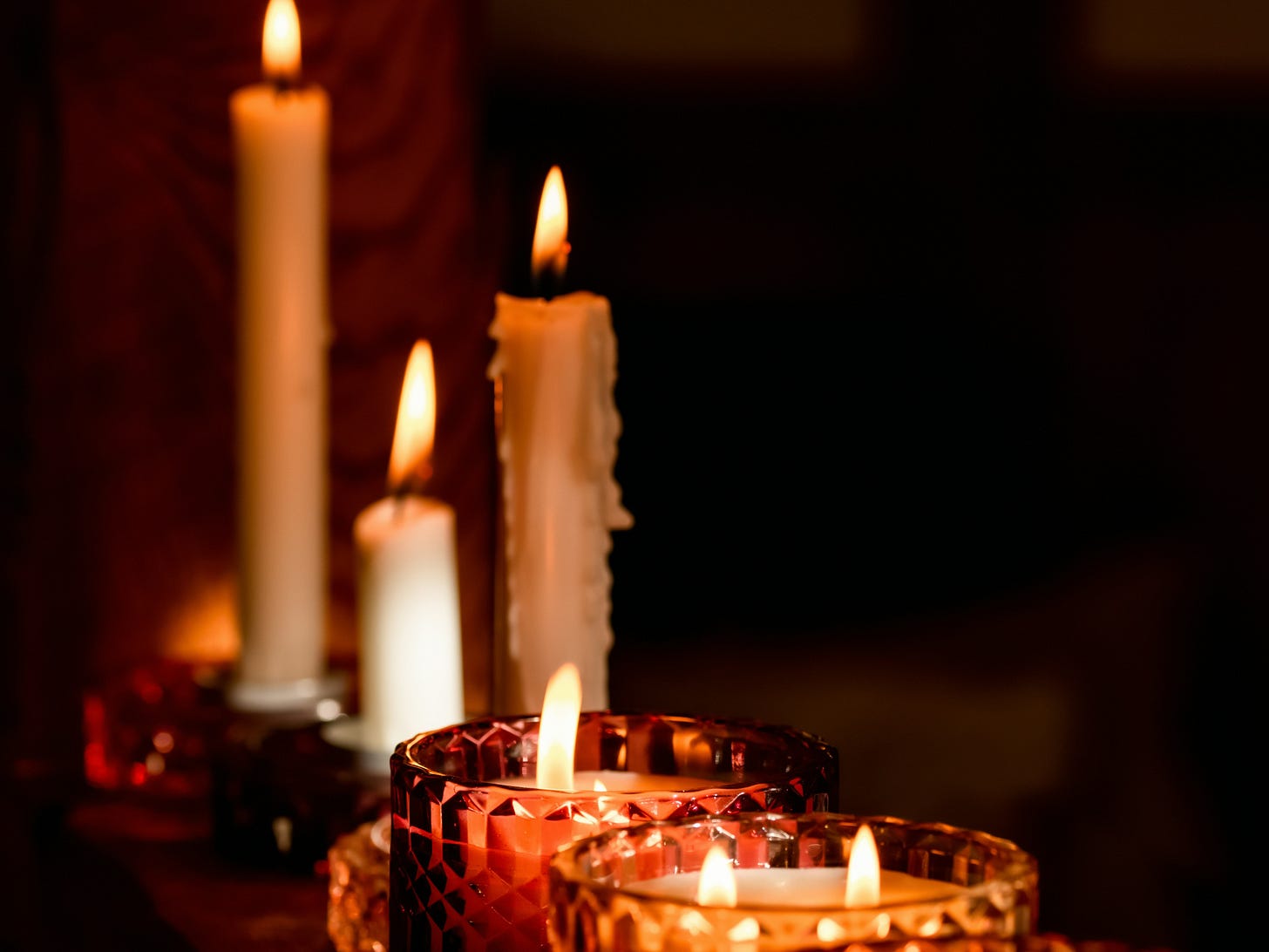 A row of lit candles in warm amber and red glass votives alongside tall white taper candles, arranged on a dark wooden surface against a dim, shadowy background. A row of lit candles in warm amber and red glass votives alongside tall white taper candles, arranged on a dark wooden surface against a dim, shadowy background.