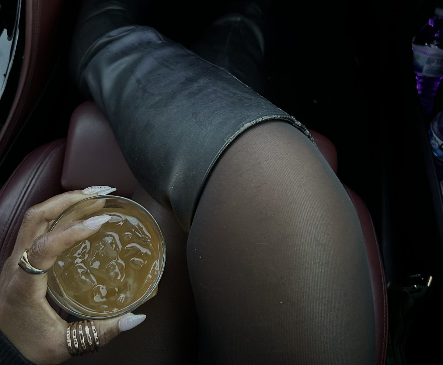 A woman seated in a car wearing black tights and knee-high leather boots holds a glass of whiskey over ice in her hand, viewed from above against a dark interior. A woman seated in a car wearing black tights and knee-high leather boots holds a glass of whiskey over ice in her hand, viewed from above against a dark interior.