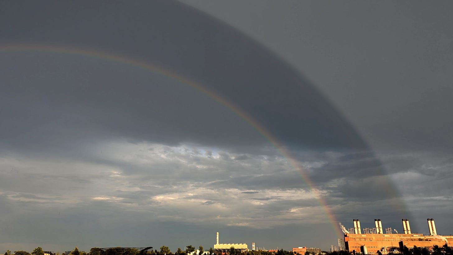 A double rainbow with a dark calling between, through a gray sky underlined by a nail clipping of earth. In the middle a white building like some kind of ship. The rainbow finds a beginning and an end with a power plant on the right side, 4 smokestacks bending in - and to be - kind. A double rainbow with a dark calling between, through a gray sky underlined by a nail clipping of earth. In the middle a white building like some kind of ship. The rainbow finds a beginning and an end with a power plant on the right side, 4 smokestacks bending in - and to be - kind.