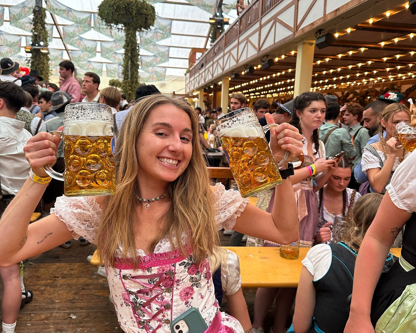 A smiling woman in a dirndl holding two large beer steins at Oktoberfest, surrounded by a crowded beer hall with long tables and festive lights. A smiling woman in a dirndl holding two large beer steins at Oktoberfest, surrounded by a crowded beer hall with long tables and festive lights.