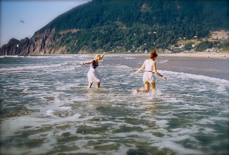 This may contain: two women are walking in the water at the beach, one is holding her arms out