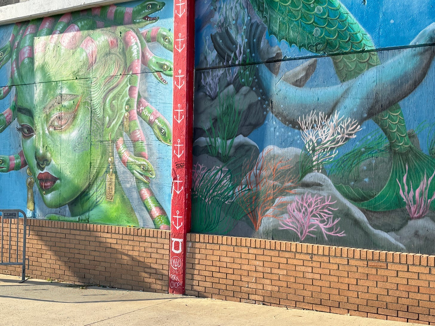 Colorful mural on a brick wall. On the left, a large green face with pink eyeshadow, gold earrings, and snakes for hair, reminiscent of Medusa. On the right, the mural transitions into an underwater scene with a mermaid’s tail, coral, seaweed, and rocks. A red vertical strip with white anchor symbols separates the two sections. Colorful mural on a brick wall. On the left, a large green face with pink eyeshadow, gold earrings, and snakes for hair, reminiscent of Medusa. On the right, the mural transitions into an underwater scene with a mermaid’s tail, coral, seaweed, and rocks. A red vertical strip with white anchor symbols separates the two sections.