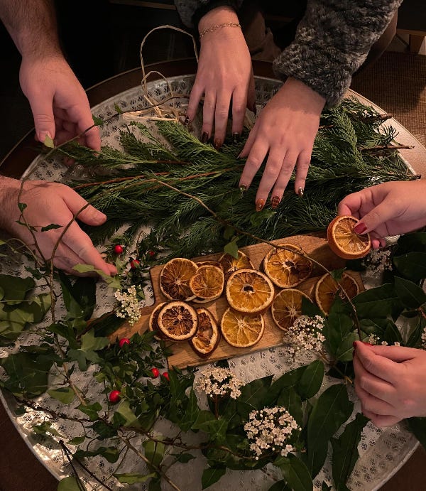 Photo d'un plateau recouvert de branches et de feuilles, et d'oranges séchées avec 6 mains qui viennent piocher ces éléments