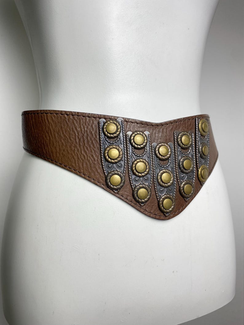 May include: A brown leather belt with decorative metal accents. The belt features a series of silver and gold-toned metal embellishments. The belt is displayed on a mannequin, showcasing its shape and design.