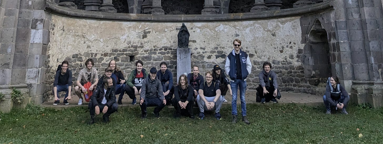 Attendees taking a group picture in the ruins of a former monastery, many are squatting with their heels down Attendees taking a group picture in the ruins of a former monastery, many are squatting with their heels down