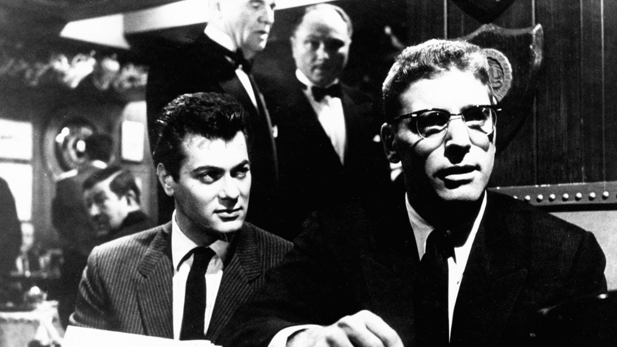 Post cover art Black-and-white scene from a film: a serious man in glasses sits at a table while another man in a suit looks at him; waiters in tuxedos stand behind them.