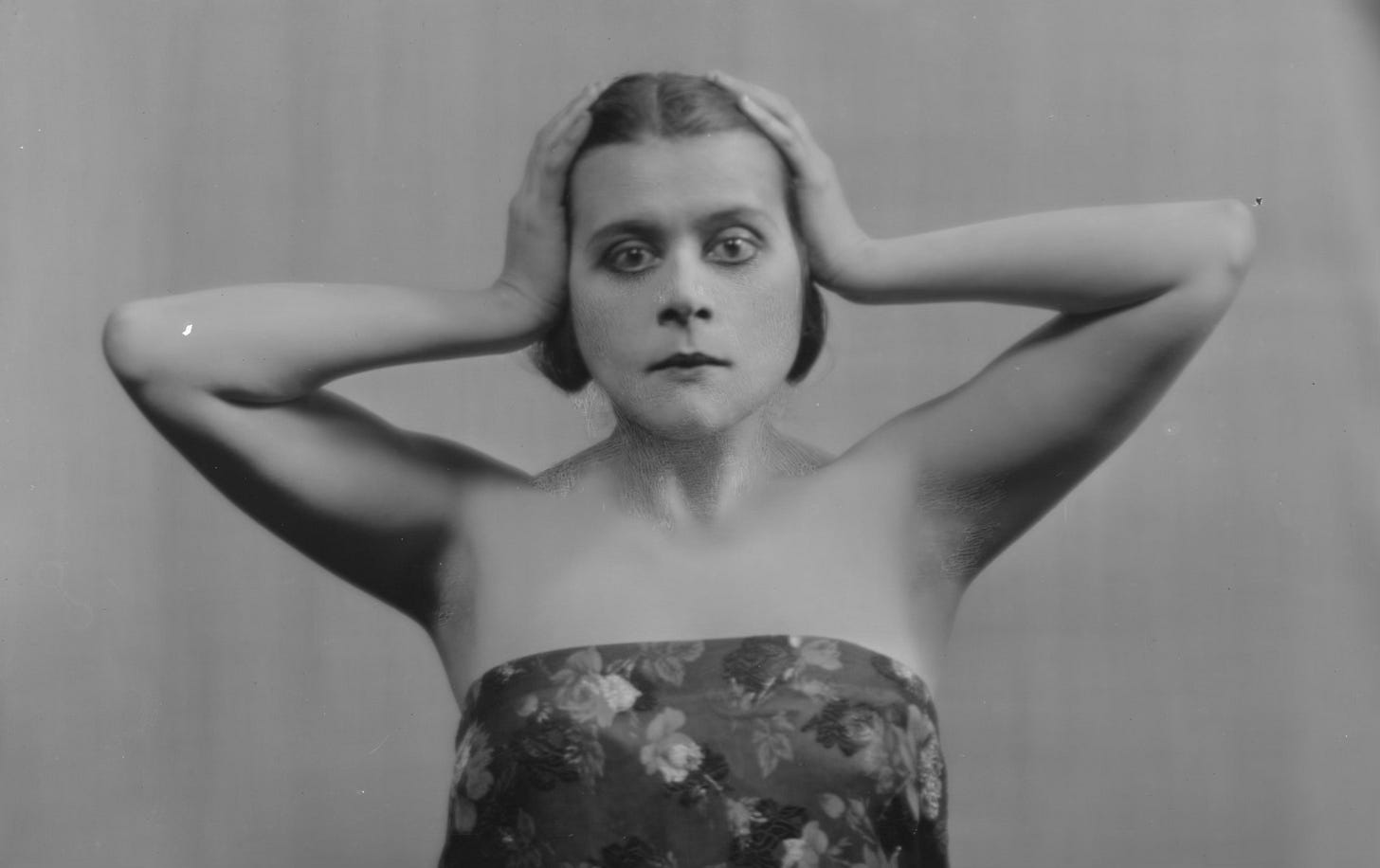Publicity photo of Theda Bara facing her camera. Her hands are pressed to the sides of her head, and she’s only wearing a floral sheet wrapped around her torso.