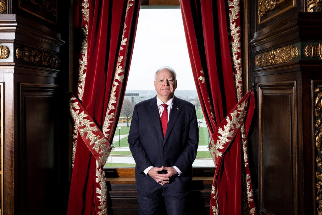 Tim Walz stands in a suit in front of a tall window clad in red velvet curtains.