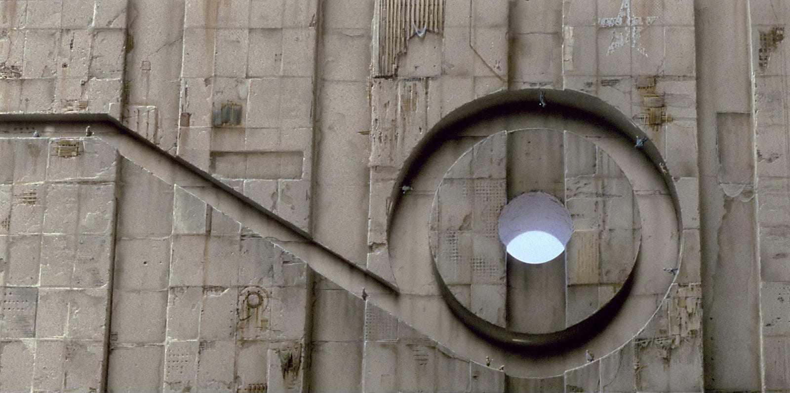 Detail from ORBIS SCIENTIARUM. Tiny figures, each carrying a light, walk a path cut into the face of an enormous textured stone facade. The path angles down before opening to a round circuit where people seem to be defying gravity as they walk the curve even upside down. The channel in the stone resembles the number 9 lying face up. A hole at the center of the loop exposes the sky outside. A boy points his light up toward it but there is no apparent path there.