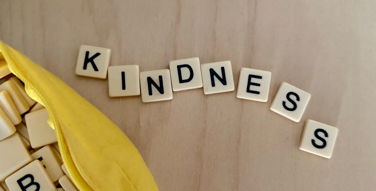 Game tiles on a light wood surface spell out KINDNESS. In the lower right corner we can see yellow bag with additional letter tiles in it.