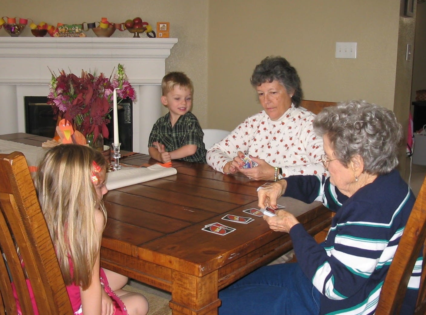 Four people sit at a wood dining room table: a 4-year-old boy with blond hair in a green plaid short-sleeve dress shirt, a woman in her 60s with salt-and-pepper hair wearing an white turtleneck with a pattern of small pumpkins, a women in her 80s, grey hair, wearing a blue and white striped sweater - she is dealing UNO cards for four people, a 7-year-old girl with long blonde hair wearing a dark pink tank top and skirt. In the background is a fire place, handmade construction paper ring chain in yellow, orange, brown draped over decorative clay and wooden bowls. On the table are two candle sticks, flowers, and a child's construction paper turkey.