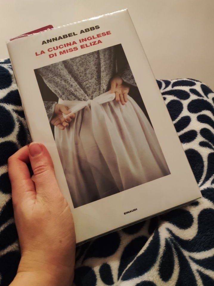 Una mano tiene un libro. Sullo sfondo una coperta di pile bianca e blu. Il libro è "La cucina inglese di Miss Eliza" e la copertina è bianca con la fotografia di una donna che si allaccia un grembiule, vista di schiena