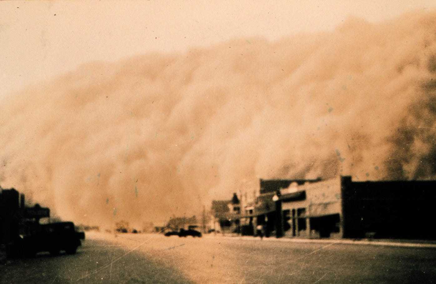 In sepia tones, a cloud of dust extending from ground to about a hundred feet above ground encroaches on a small town, with 1930s-era vehicles parked in the street. In sepia tones, a cloud of dust extending from ground to about a hundred feet above ground encroaches on a small town, with 1930s-era vehicles parked in the street.