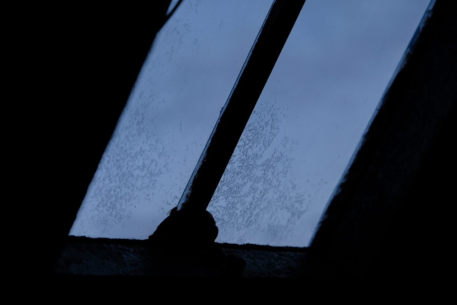 Fading sunlight filters through a cracked, rust-stained window inside a bothy, casting the room into deepening shadow as night approaches.