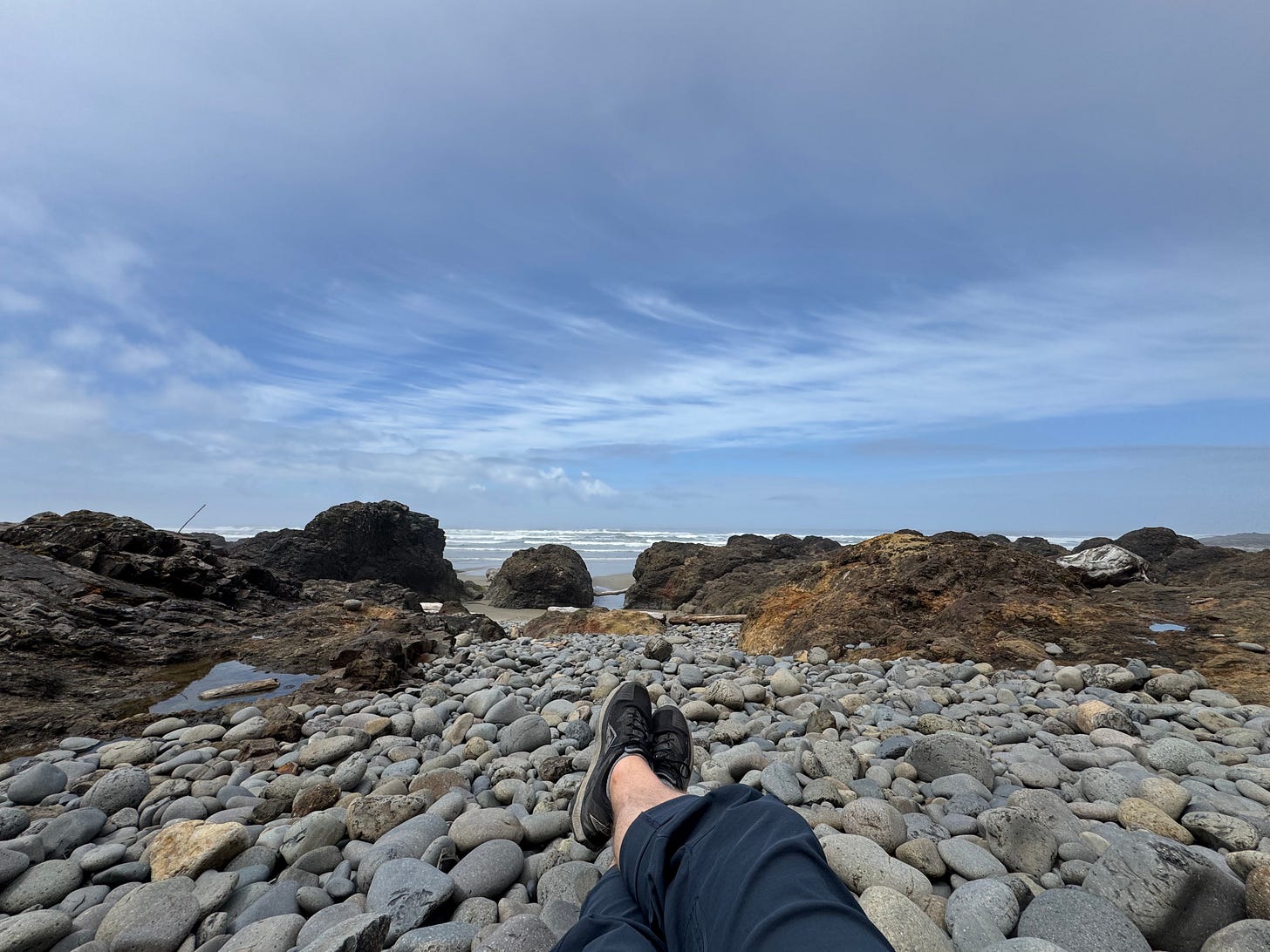 First-person view of legs and feet in black shoes sitting on smooth river stones at Neptune Beach, Oregon, with rocky tide pools and Pacific Ocean waves in the background under an overcast sky