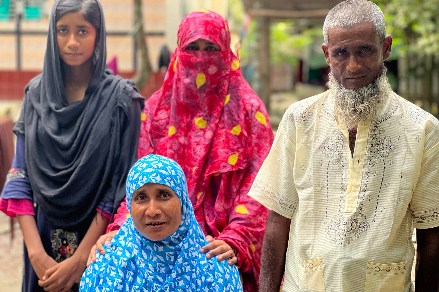 Rahima Banu, seated in front of her husband and two daughters, wears a cobalt-blue scarf with white flowers — it’s draped over her head and shoulders and modestly tucked under her chin. The daughter who stands immediately behind her rests her hands on her mother’s shoulders; she is draped in bright pink cloth that covers all but her eyes. The daughter to the viewer’s left wears a black scarf that half covers her head, and she holds her hands together in front of her. Her husband is in a daffodil-yellow short-sleeved shirt, his gaze fixed on the camera in front of him.