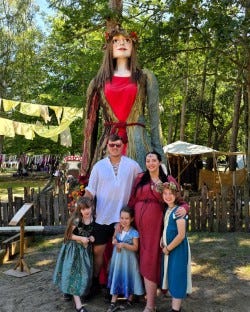 The Barker/Marney family of a tall man, woman and 3 girls plus a bump. a;ll dressed in medieval clothing standing in front of Flora, The Singleton Giant (wicker framed giant lady) sited in a woodland setting with bunting and tents for a festival.