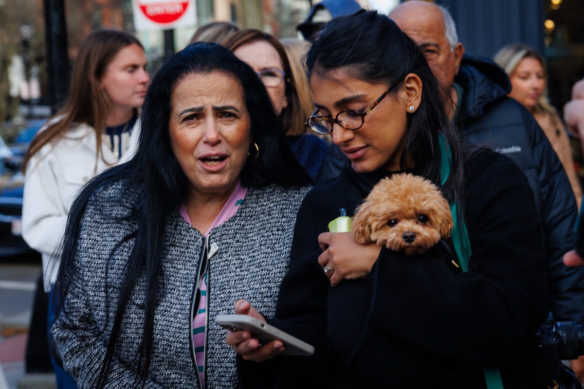 Two women stand in a busy crowd; one reacts toward the camera while the other holds a small fluffy dog.