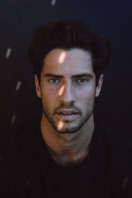Free Handsome young pensive Hispanic male model with dark hair in casual clothes looking at camera with concentration in black studio with sun flares Stock Photo