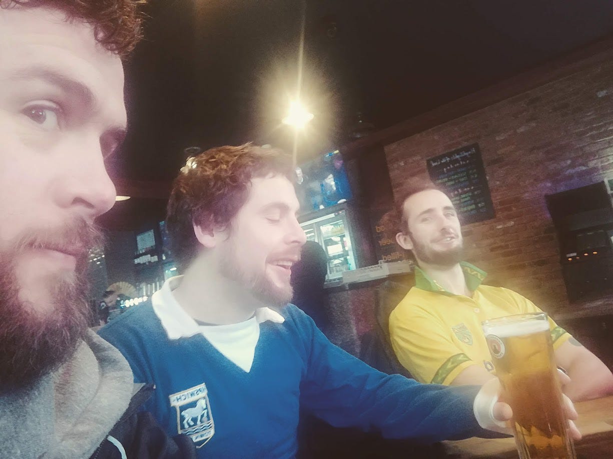 Three men in their early thirties are sat in a pub. The picture is a selfie by one of them. They are say around a table. One wears a retro ipswich shirt, one a norwich shirt. The other is neutral. 