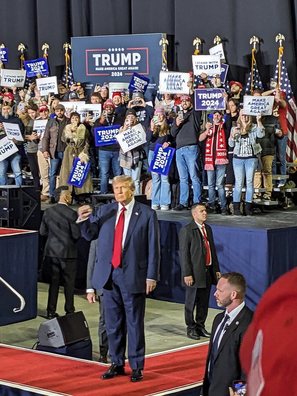 Trump stands in front of people and flags, looking rumpled and low energy and making a fist Trump stands in front of people and flags, looking rumpled and low energy and making a fist