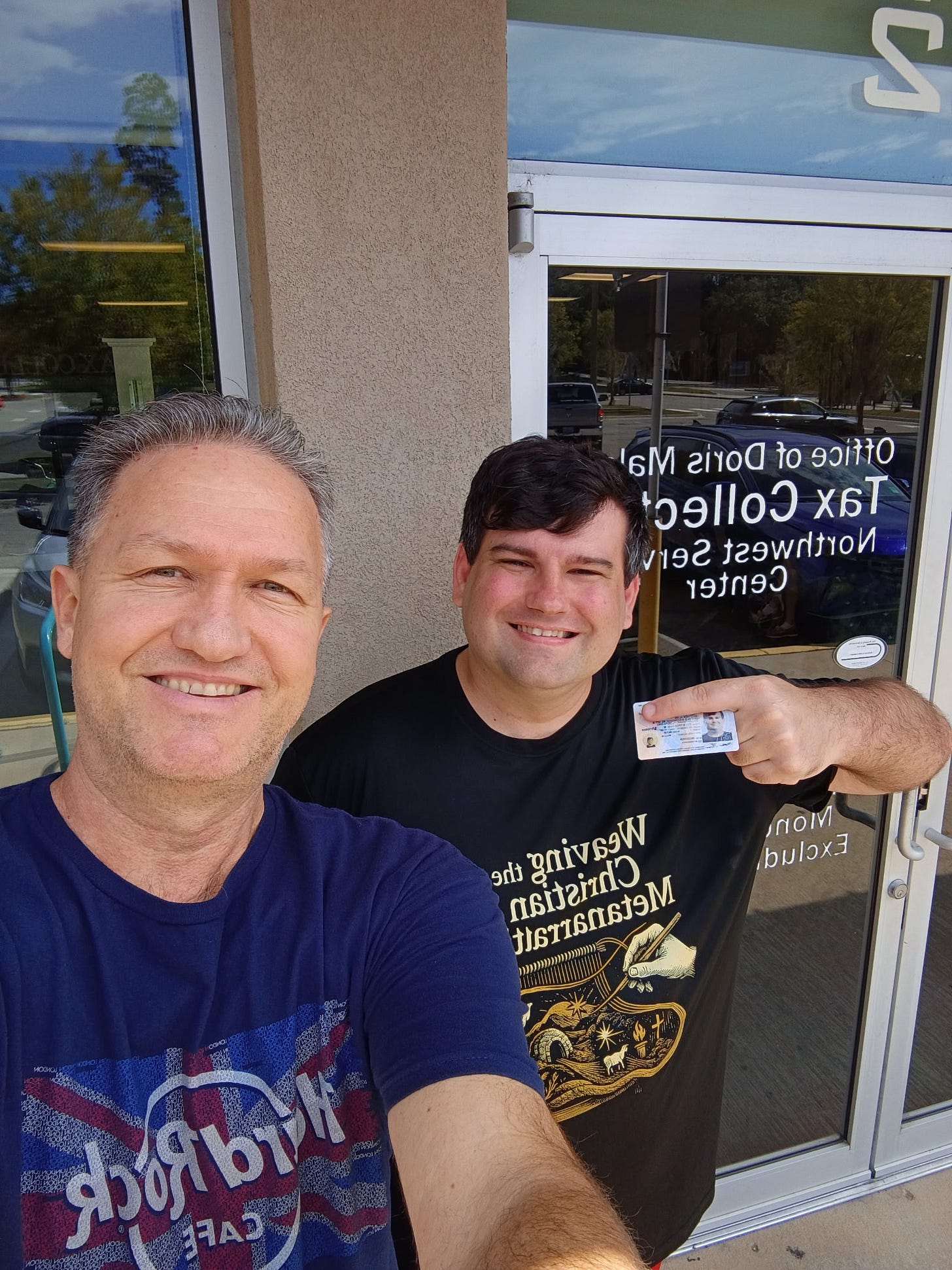 A selfie of two smiling men standing outside a storefront; the man on the right points to an ID card. Behind them is a glass door with white lettering reading “Office of Doris Mal… Tax Collect… Northwest Serv… Center,” reflecting parked cars and trees, and a large “2” sign above the entrance.